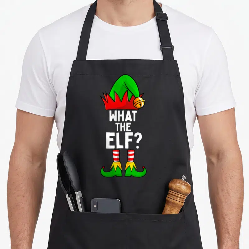 Cute family christmas elf apron - What The Elf pun with elf hat, striped socks, and green shoes — shown on a black cotton canvas apron against a white background