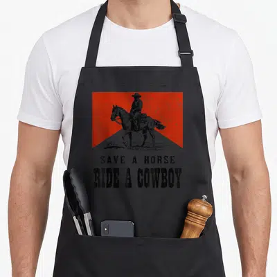 Country cowboy apron - Distressed SAVE A HORSE RIDE A COWBOY text below black cowboy silhouette on red background — shown on a black cotton canvas apron against a white background
