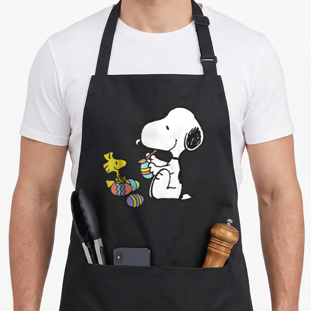 Woodstock easter apron - Snoopy painting striped egg next to Woodstock and three decorated eggs — shown on a black cotton canvas apron against a white background