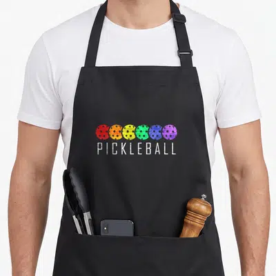 Paddle pickleball apron - Six colorful pickleballs in red orange yellow green blue purple above white PICKLEBALL text — shown on a black cotton canvas apron against a white background