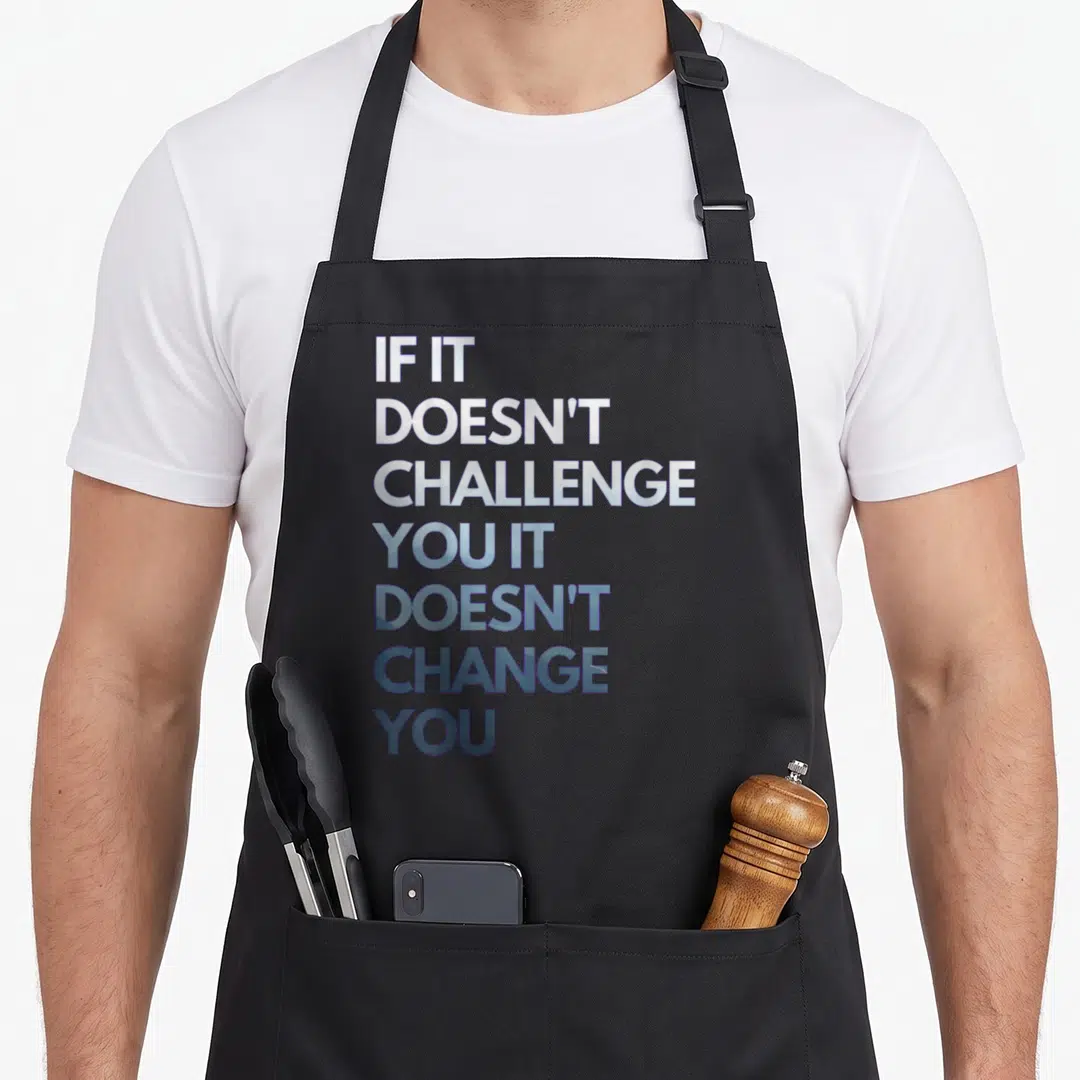 Fitness motivation apron - Stacked white and blue text reading IF IT DOESN'T CHALLENGE YOU IT DOESN'T CHANGE YOU — shown on a black cotton canvas apron against a white background