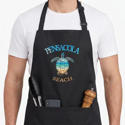 Jcomb pensacola apron - Sea turtle with beach landscape shell and arched PENSACOLA BEACH text in blue and orange — shown on a black cotton canvas apron against a white background