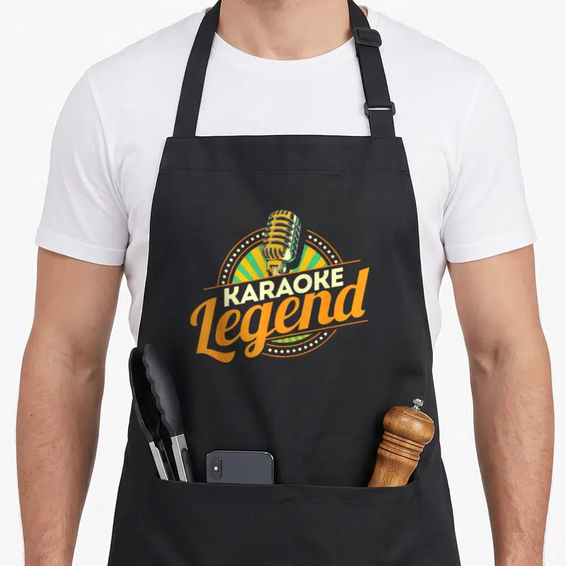 Legend karaoke apron - Vintage microphone over green orange sunburst with KARAOKE Legend text — shown on a black cotton canvas apron against a white background