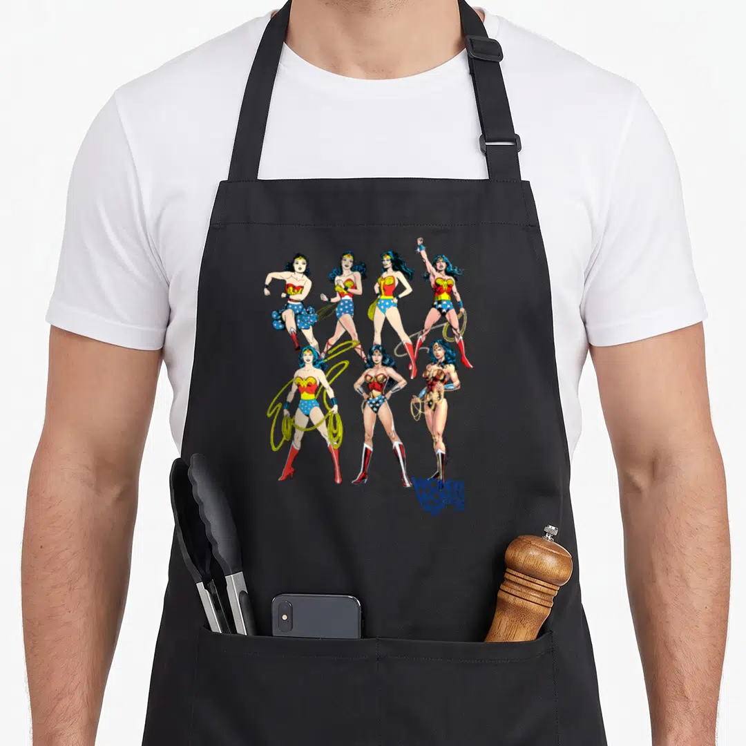 Decade curiosity apron - Seven Wonder Woman illustrations in blue red and gold with WONDER WOMAN 75 text at bottom right — shown on a black cotton canvas apron against a white background