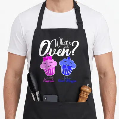 Oven muffin man apron - White script text above pink and blue anthropomorphic cupcakes with bows — shown on a black cotton canvas apron against a white background