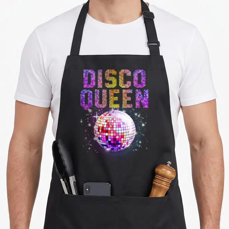 Disco queen apron - Stacked DISCO QUEEN text above a multicolored disco ball with white sparkles on purple background — shown on a black cotton canvas apron against a white background