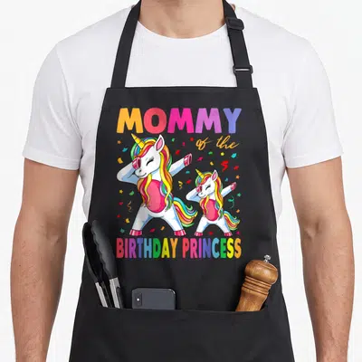 Dabbing unicorn princess apron - Two white unicorns with rainbow manes dabbing below MOMMY of the BIRTHDAY PRINCESS — shown on a black cotton canvas apron against a white background