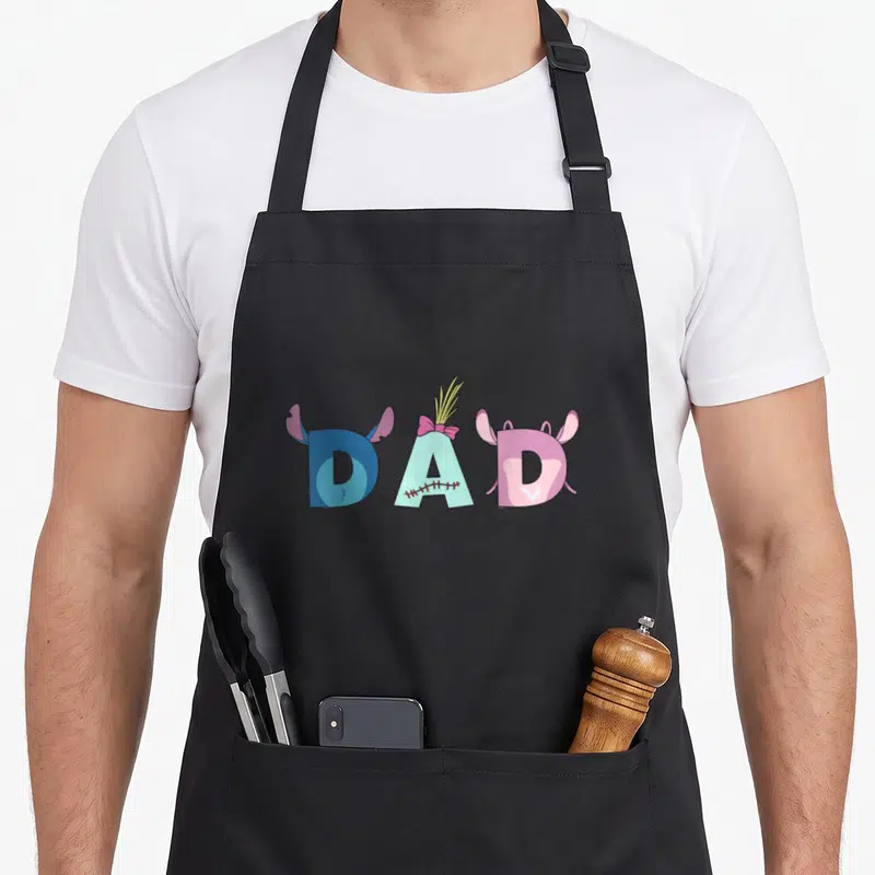 Trip father day apron - Cartoon DAD text with blue D, stitched A with pink bow, and pink D with purple ears — shown on a black cotton canvas apron against a white background