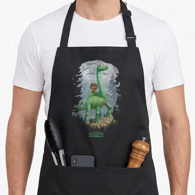 Arlo dinosaur apron - Green dinosaur stands on brown rock with boy on back, blue-grey trees, pterodactyls, THE GOOD — shown on a black cotton canvas apron against a white background