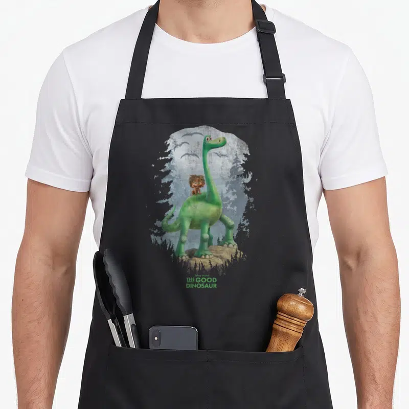 Arlo dinosaur apron - Green dinosaur stands on brown rock with boy on back, blue-grey trees, pterodactyls, THE GOOD — shown on a black cotton canvas apron against a white background