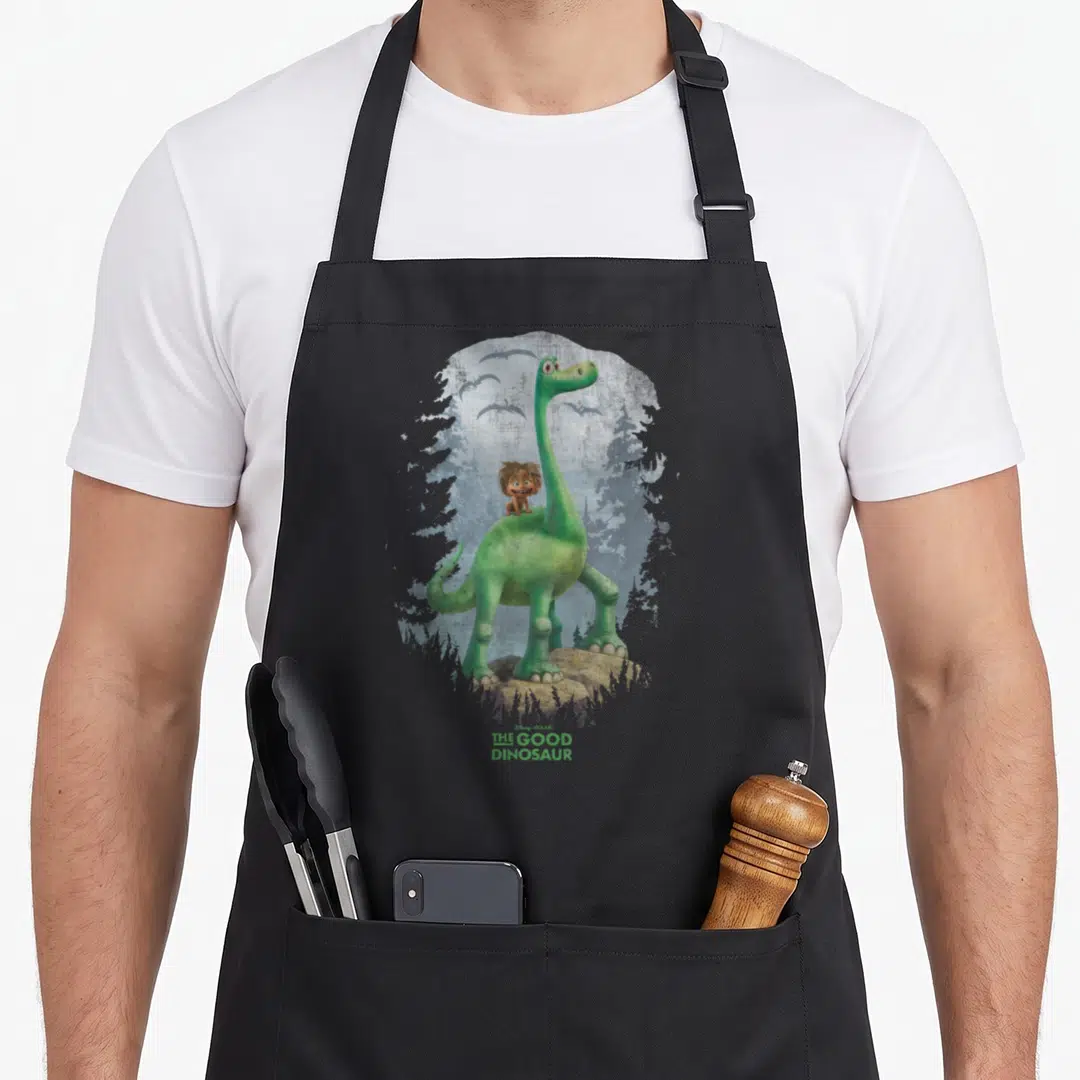Arlo dinosaur apron - Green dinosaur stands on brown rock with boy on back, blue-grey trees, pterodactyls, THE GOOD — shown on a black cotton canvas apron against a white background