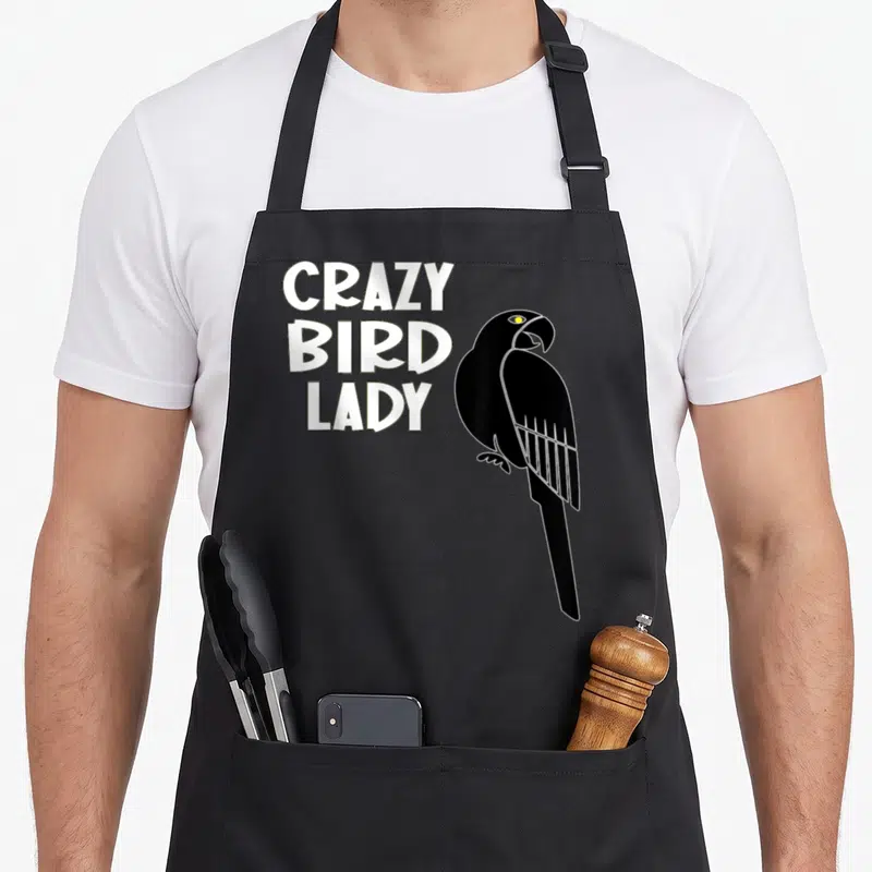 Birdie bird apron - White stacked text CRAZY BIRD LADY beside a black cartoon parrot with a yellow eye — shown on a black cotton canvas apron against a white background