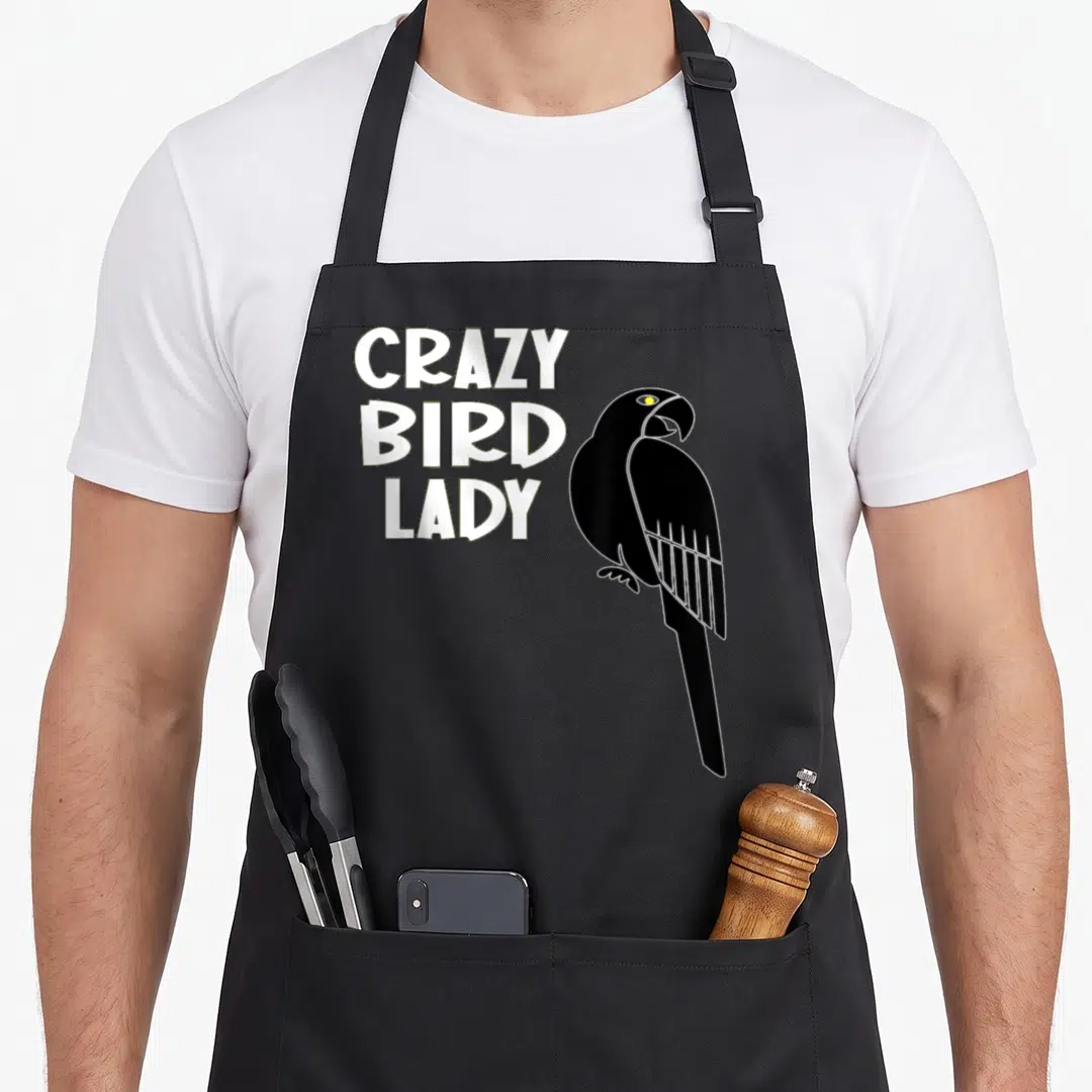 Birdie bird apron - White stacked text CRAZY BIRD LADY beside a black cartoon parrot with a yellow eye — shown on a black cotton canvas apron against a white background