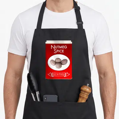 Nutmeg spice apron - Red spice tin graphic with NUTMEG SPICE text and four brown nutmeg seeds in a white oval — shown on a black cotton canvas apron against a white background