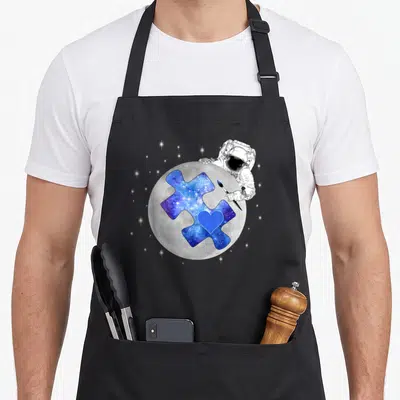 Space autism apron - Astronaut painting blue galaxy puzzle piece with heart on grey moon — shown on a black cotton canvas apron against a white background