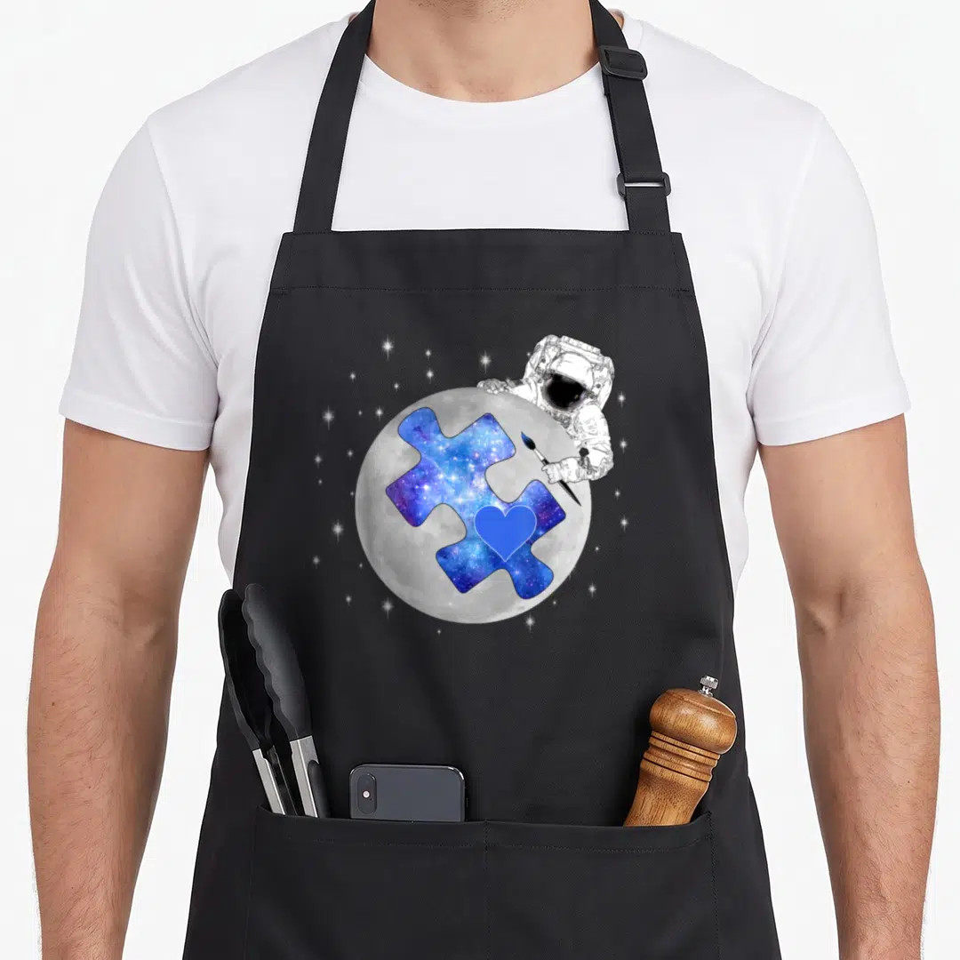 Space autism apron - Astronaut painting blue galaxy puzzle piece with heart on grey moon — shown on a black cotton canvas apron against a white background