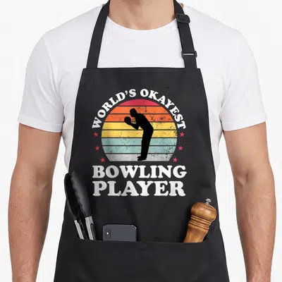 Okayest bowling lover apron - Retro sun with bowler silhouette and WORLD'S OKAYEST BOWLING PLAYER text in white, red — shown on a black cotton canvas apron against a white background