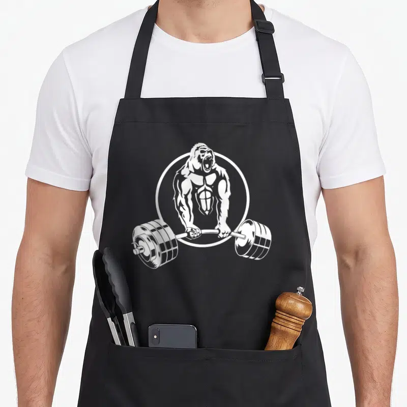 Strength gorilla mom apron - White line art of a muscular gorilla holding a barbell inside a circle on an apron — shown on a black cotton canvas apron against a white background