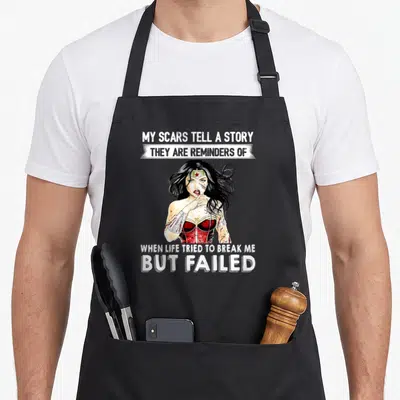 Reminder the story of the apron - Realistic Wonder Woman with blood and white text reading My Scars Tell A Story — shown on a black cotton canvas apron against a white background