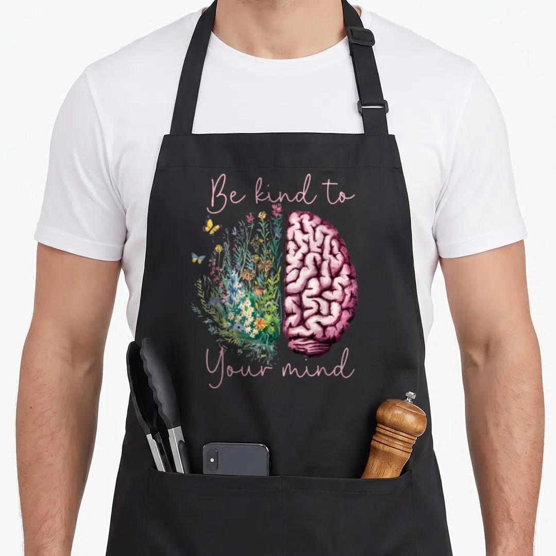 Mind anxiety awareness apron - Split brain illustration with flowers and butterflies and pink text reading Be kind — shown on a black cotton canvas apron against a white background