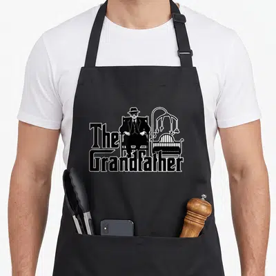 Grandfather grandad aprons - The Grandfather text with suited man and baby crib illustration — shown on a black cotton canvas apron against a white background