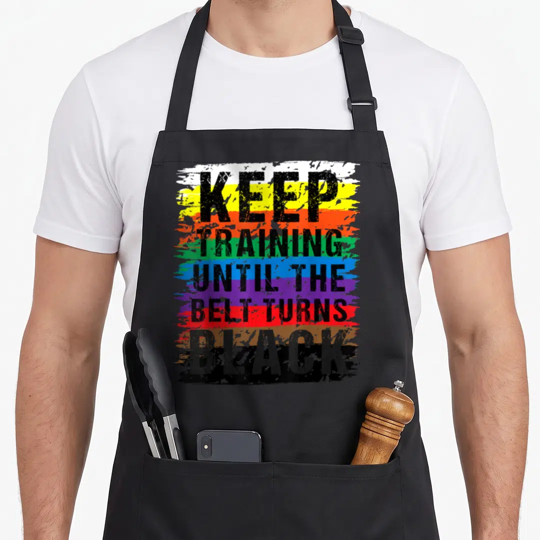 Belt i turn grills on apron - Stacked black text reading Keep Training Until The Belt Turns Black over horizontal — shown on a black cotton canvas apron against a white background