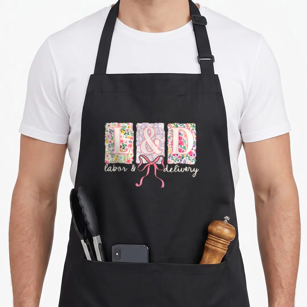 Spring nurse apron - L and D floral blocks with pink bow and script text on black apron — shown on a black cotton canvas apron against a white background