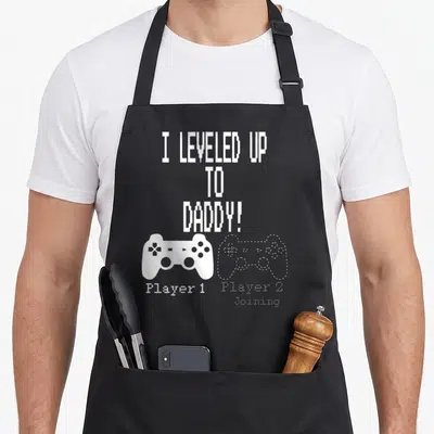 Parent daddy apron - White pixel text I leveled up to daddy with solid and outlined game controllers side by side — shown on a black cotton canvas apron against a white background