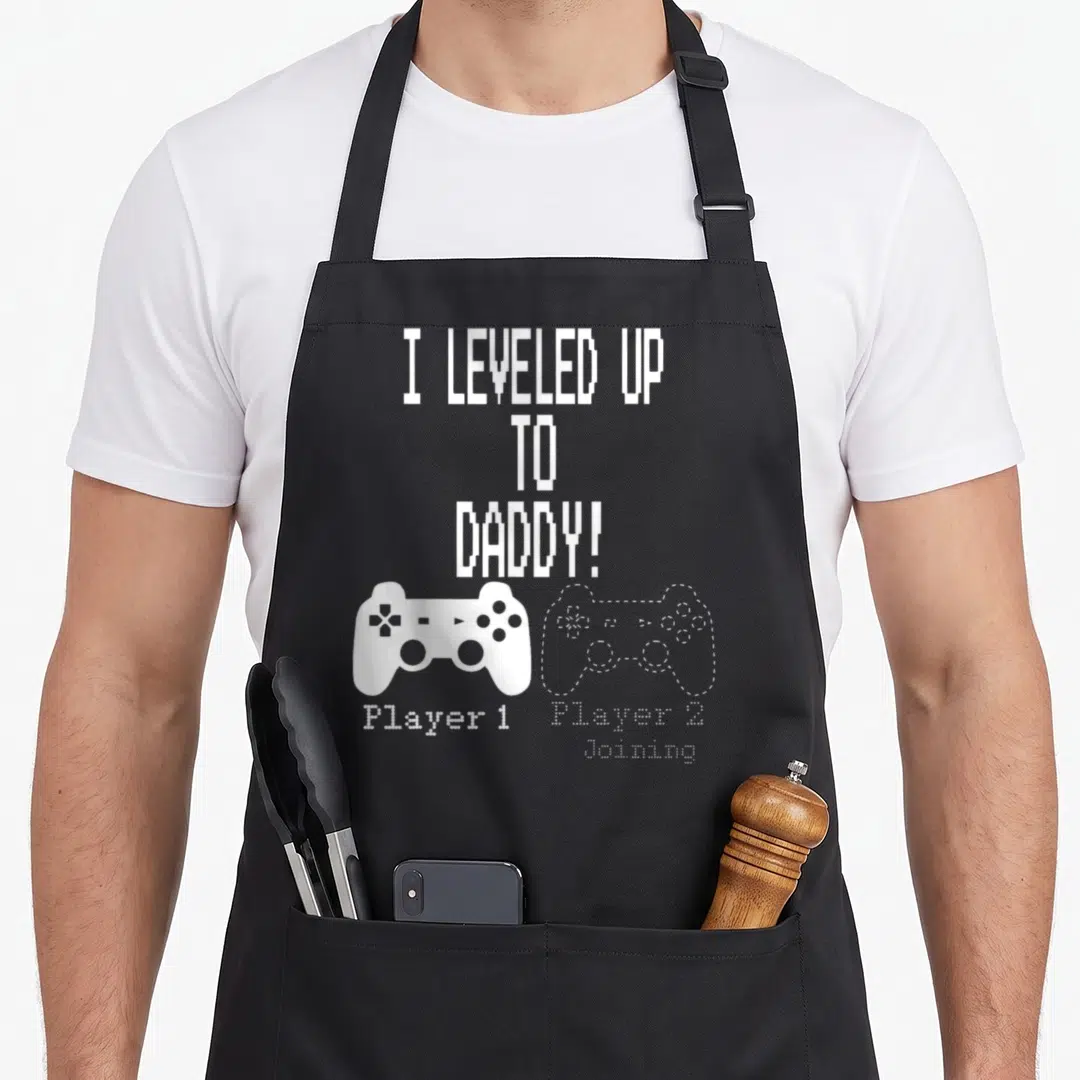 Parent daddy apron - White pixel text I leveled up to daddy with solid and outlined game controllers side by side — shown on a black cotton canvas apron against a white background