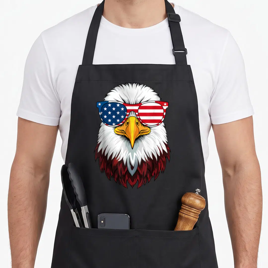 4th eagles grill apron - Cartoon bald eagle head wearing sunglasses with blue stars and red white stripes — shown on a black cotton canvas apron against a white background