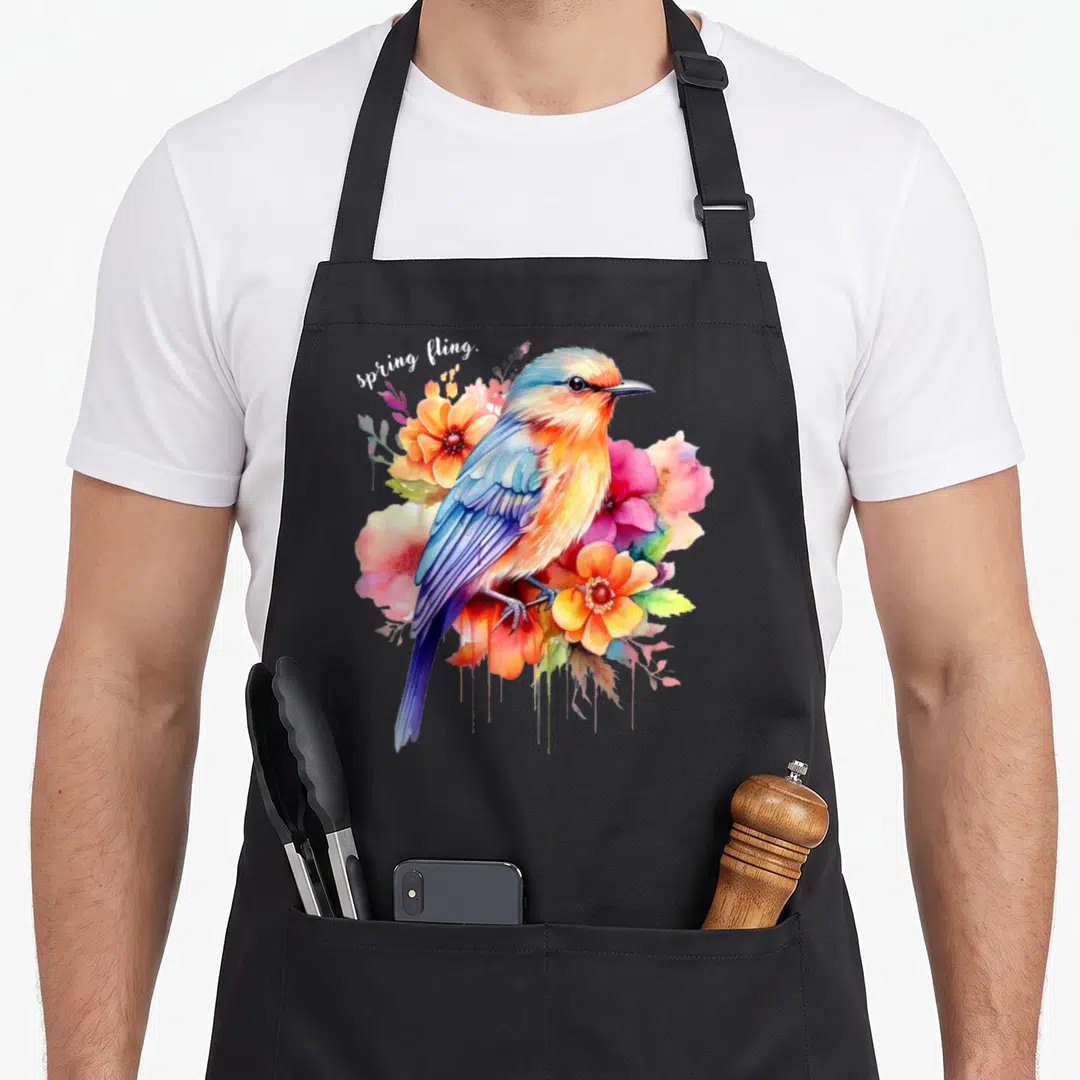 Spring the floral apron - Watercolor bird in blue and orange surrounded by pink flowers with spring fling text — shown on a black cotton canvas apron against a white background