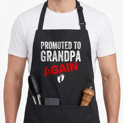 Pregnancy grandpa apron - Distressed white and red text Promoted to Grandpa Again with two white baby footprints below — shown on a black cotton canvas apron against a white background