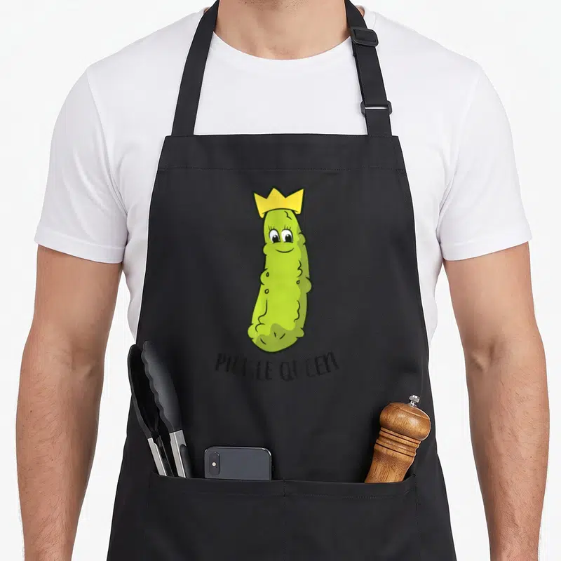 Queen pickle apron - Cartoon green pickle wearing a yellow crown above black PICKLE QUEEN text — shown on a black cotton canvas apron against a white background