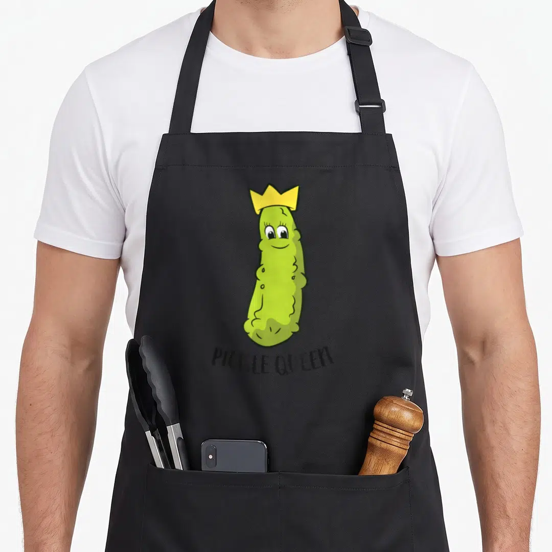 Queen pickle apron - Cartoon green pickle wearing a yellow crown above black PICKLE QUEEN text — shown on a black cotton canvas apron against a white background