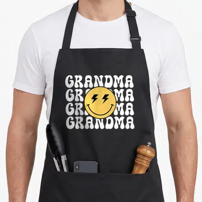 Grandma custom face apron - Yellow smiley with lightning eyes over four stacked white GRANDMA words on black background — shown on a black cotton canvas apron against a white background