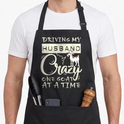 Husband driving aprons - Cartoon goat standing next to text DRIVING MY HUSBAND Crazy ONE GOAT AT A TIME — shown on a black cotton canvas apron against a white background