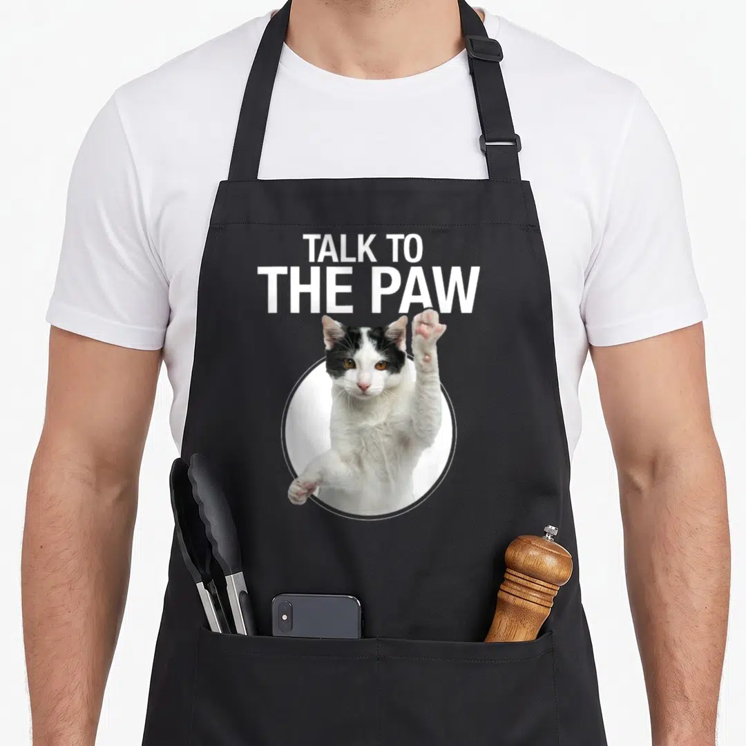 Talking cat apron - TALK TO THE PAW text above black and white cat raising paw in white circle — shown on a black cotton canvas apron against a white background