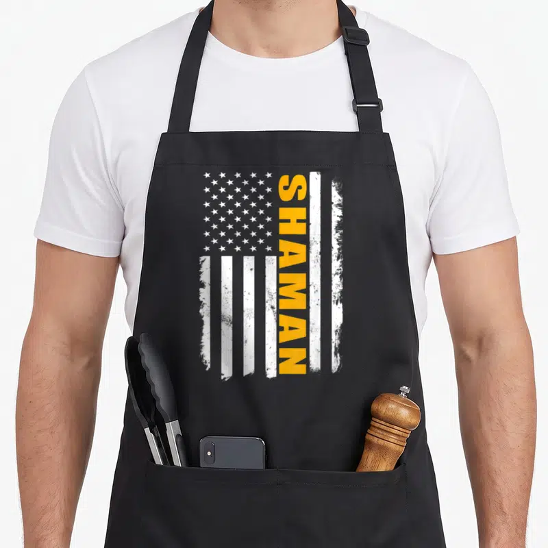 Shaman american flag apron - Vertical orange SHAMAN text overlaying distressed white stripes and stars on grey — shown on a black cotton canvas apron against a white background