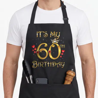 60th queen apron - Gold IT'S MY 60TH BIRTHDAY text with crown, red hearts, and diamond-embedded letters — shown on a black cotton canvas apron against a white background