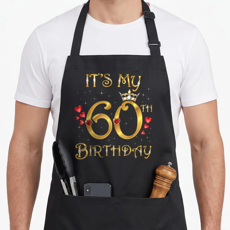 60th queen apron - Gold IT'S MY 60TH BIRTHDAY text with crown, red hearts, and diamond-embedded letters — shown on a black cotton canvas apron against a white background