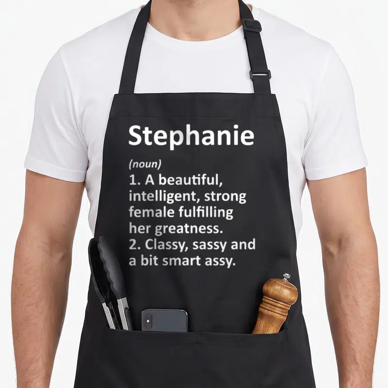 Stephanie christmas apron - White text defining Stephanie as a beautiful, intelligent, strong, and sassy noun — shown on a black cotton canvas apron against a white background