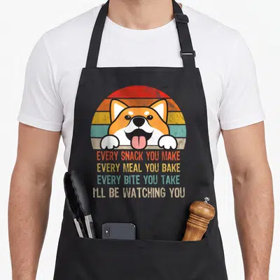 Shiba junkie apron - Retro shiba inu peeking over striped sunset with four lines of text about watching snacks and meals — shown on a black cotton canvas apron against a white background
