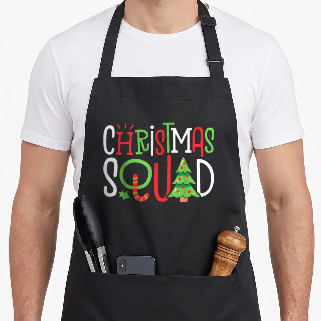 Squad christmas tree apron - CHRISTMAS SQUAD text with cartoon tree, candy cane, and holly leaf in red, green, and white — shown on a black cotton canvas apron against a white background