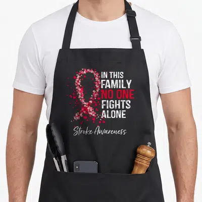 Stroke family aprons - Heart-shaped awareness ribbon and stacked text IN THIS FAMILY NO ONE FIGHTS ALONE — shown on a black cotton canvas apron against a white background