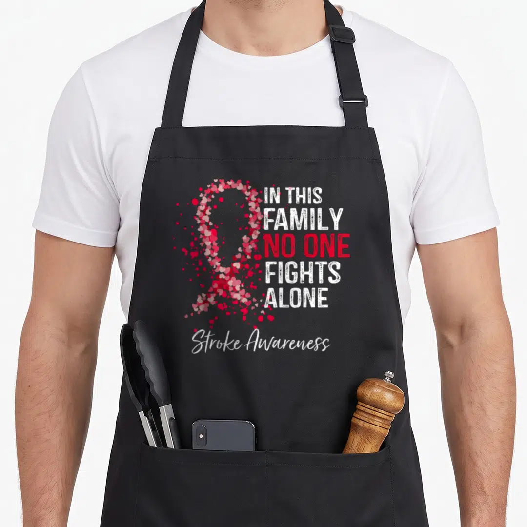 Stroke family aprons - Heart-shaped awareness ribbon and stacked text IN THIS FAMILY NO ONE FIGHTS ALONE — shown on a black cotton canvas apron against a white background