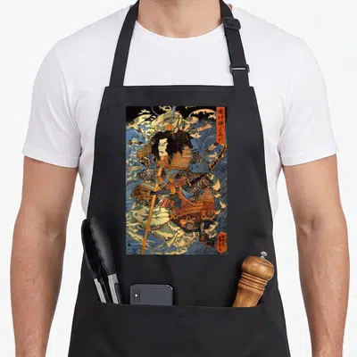 Japanese crab apron - Samurai in blue and orange armor holding a spear amid crashing waves with vertical Japanese text — shown on a black cotton canvas apron against a white background