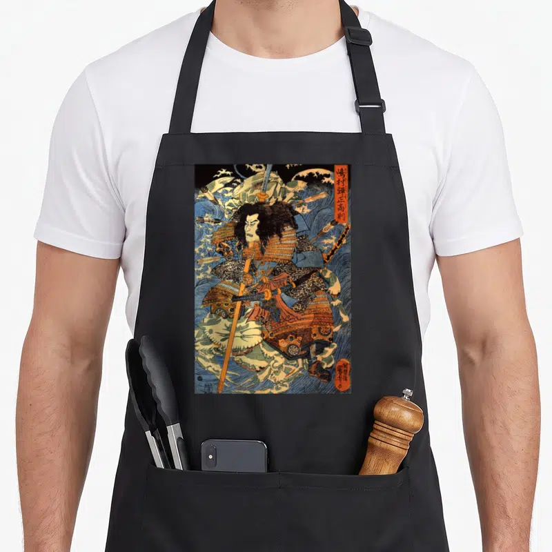 Japanese crab apron - Samurai in blue and orange armor holding a spear amid crashing waves with vertical Japanese text — shown on a black cotton canvas apron against a white background
