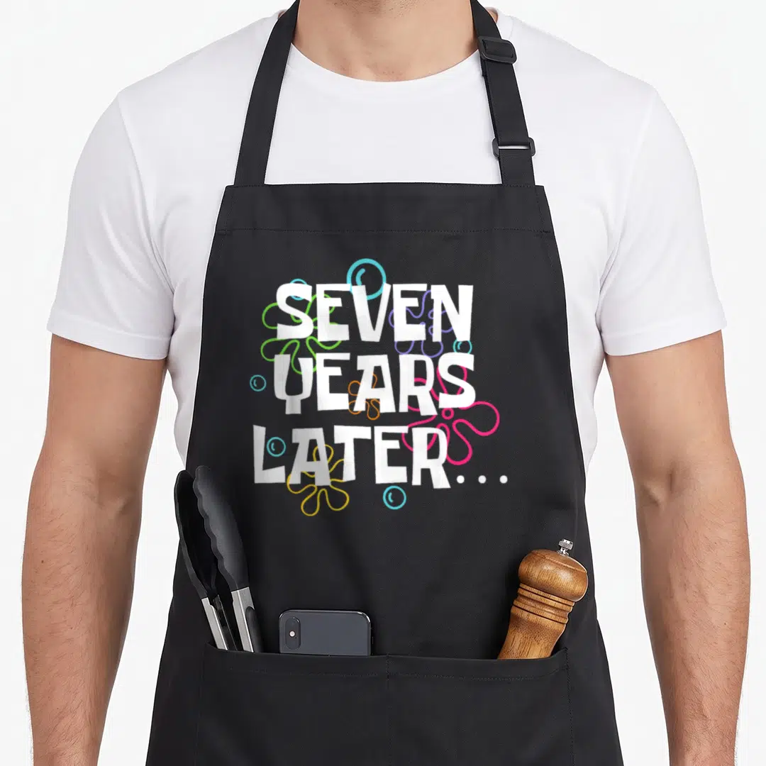 Later meme apron - Stacked white SEVEN YEARS LATER text with colorful cartoon flowers and bubbles — shown on a black cotton canvas apron against a white background