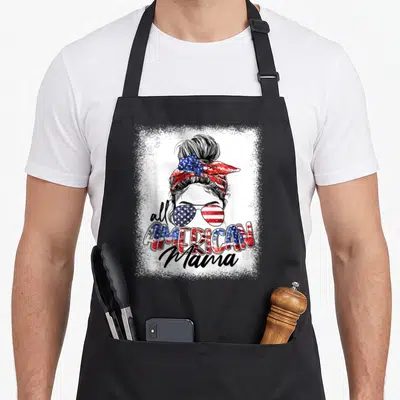 Mama bun messy aprons - Woman with messy bun, flag sunglasses, and all AMERICAN Mama text in red white blue distressed — shown on a black cotton canvas apron against a white background
