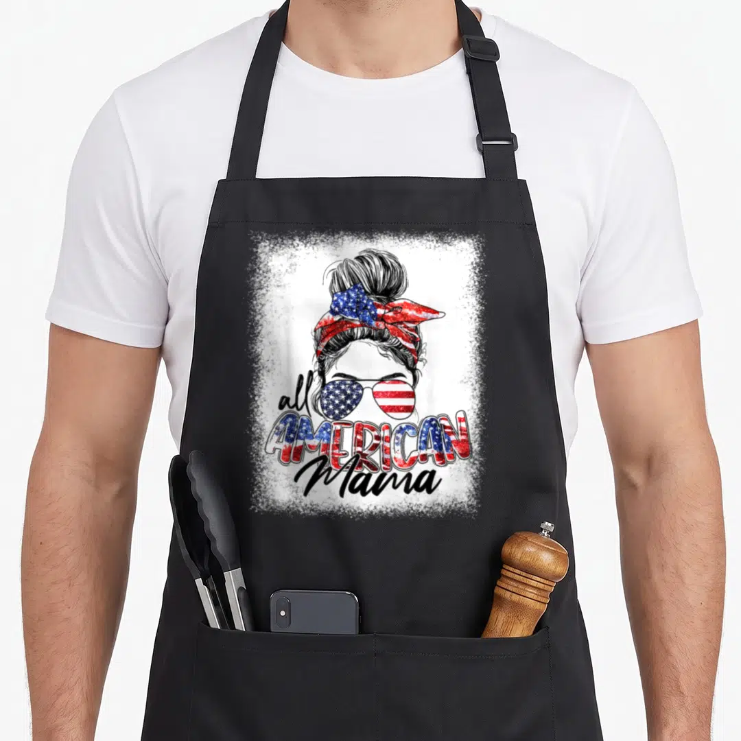 Mama bun messy aprons - Woman with messy bun, flag sunglasses, and all AMERICAN Mama text in red white blue distressed — shown on a black cotton canvas apron against a white background